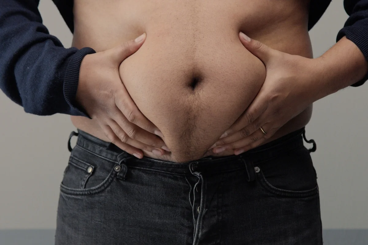 Homem com sobrepeso em close-up, vista frontal da barriga, levantando a camisa e apertando a gordura abdominal. Metrópoles